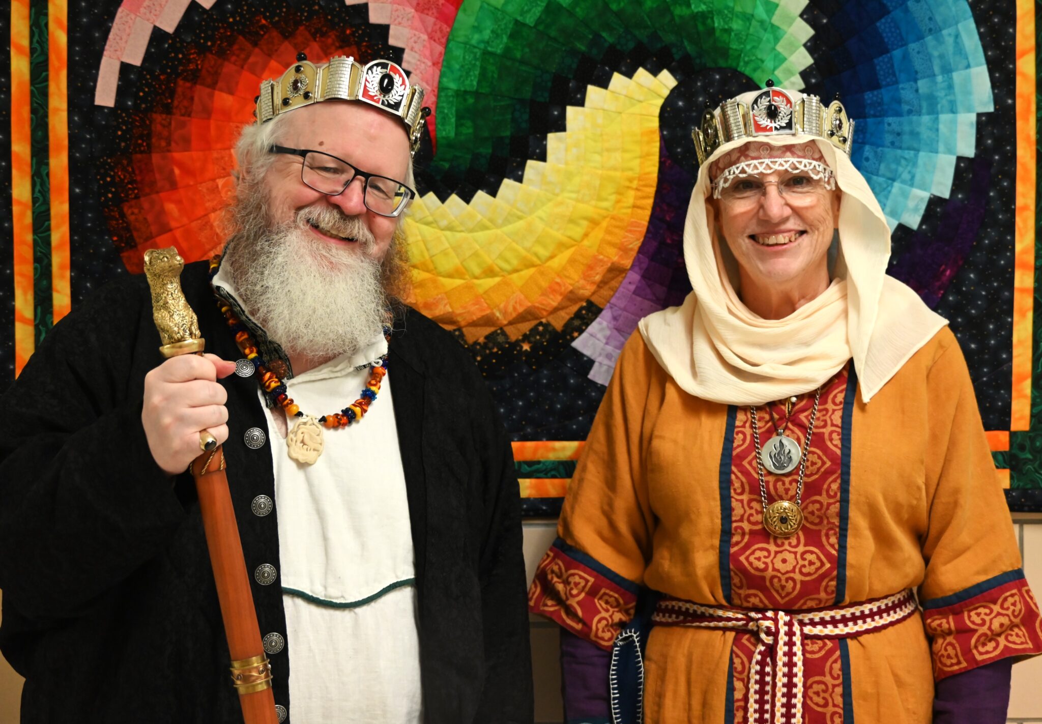Two people standing in front of a colorful patchwork quilt. On the left is Baron Nikolai, on the right is Baroness Taitiana. Both wear the Baronial crowns and respective mediallions for their awards.
